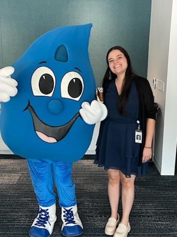 Student intern in a navy blue dress and tan shoes stands next to Charlotte Water's mascot, the bright blue waterdrop with big eyes, smile and white gloves