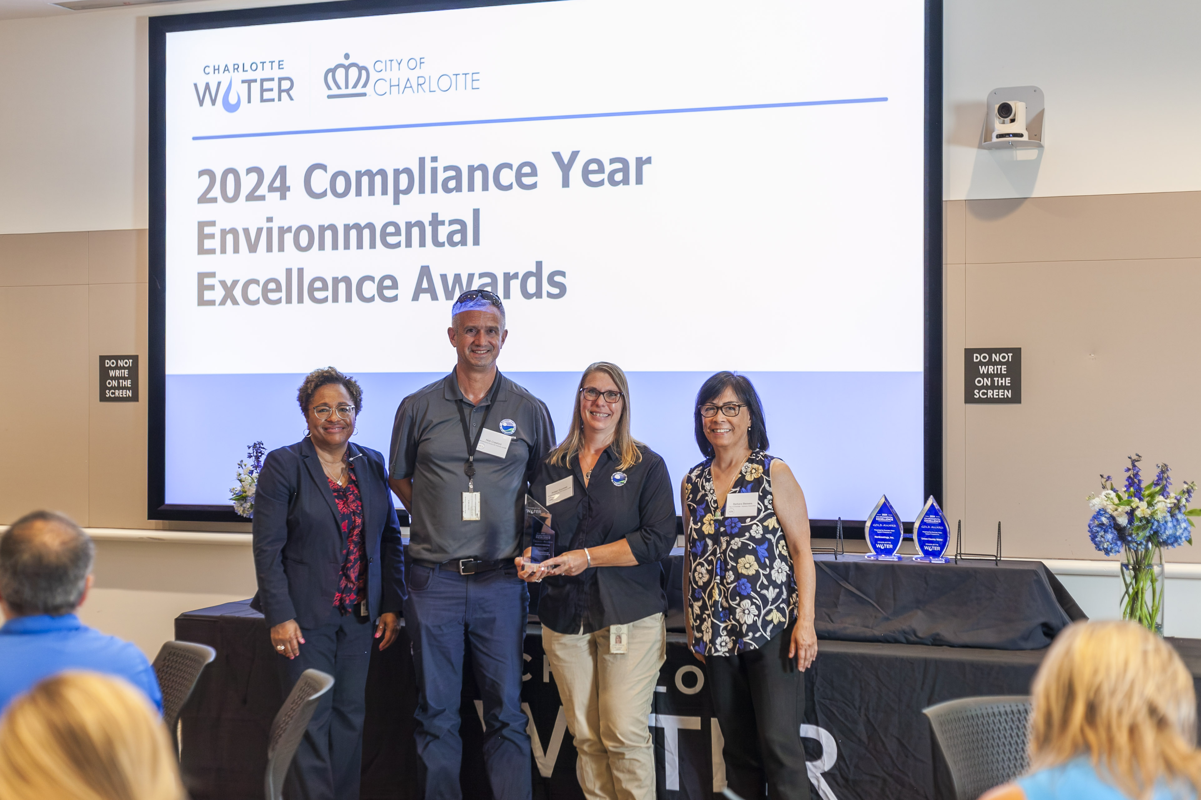4 people stand in front of a large video screen announcing 2024 compliance year environmental excellence awards for Charlotte Water and City of Charlotte, one individual holds a blue and glass trophy