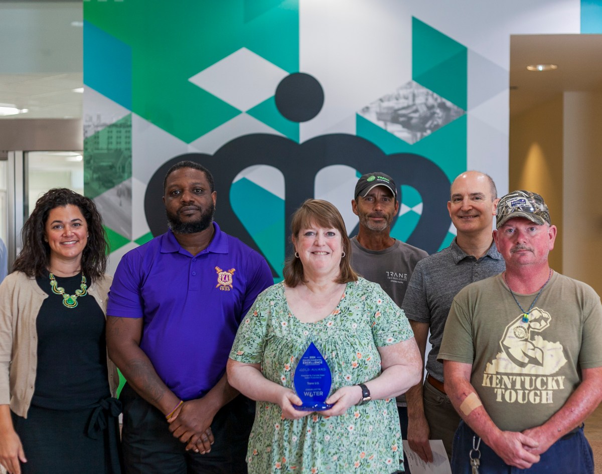 1 of 6 recipients holds a blue and glass trophy as all 6 stand in front of green and white mural featuring the city of Charlotte logo