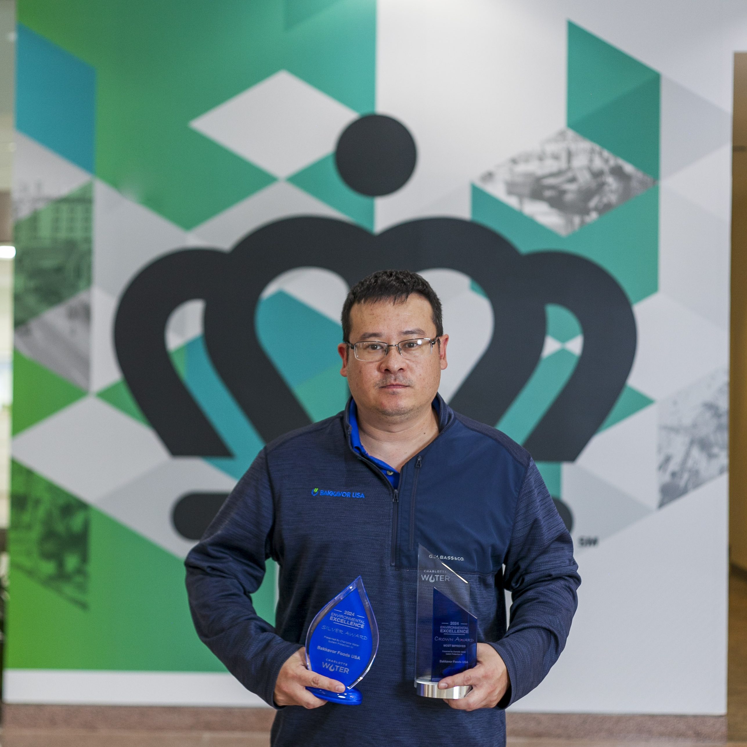 Utility employee with dark hair, glasses, blue jacket holds 2 blue and clear glass trophies in front of large green and white mural featuring the city of Charlotte logo