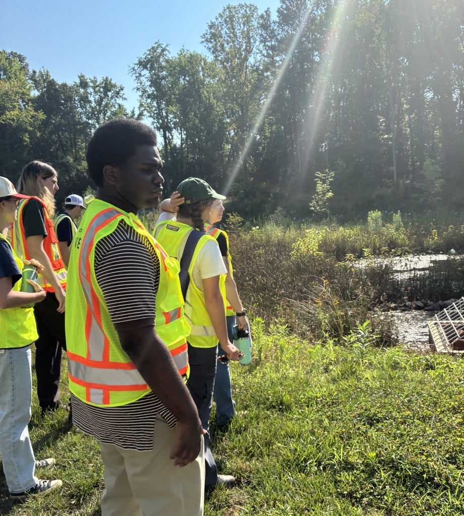 Group of student interns wearing yellow safety vests tour a water utility installation near a pond