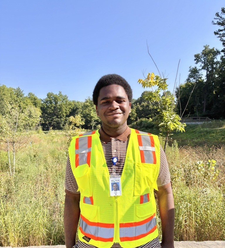 Student intern wearing yellow safety vest stands smiling outdoors in front of a grassy meadow