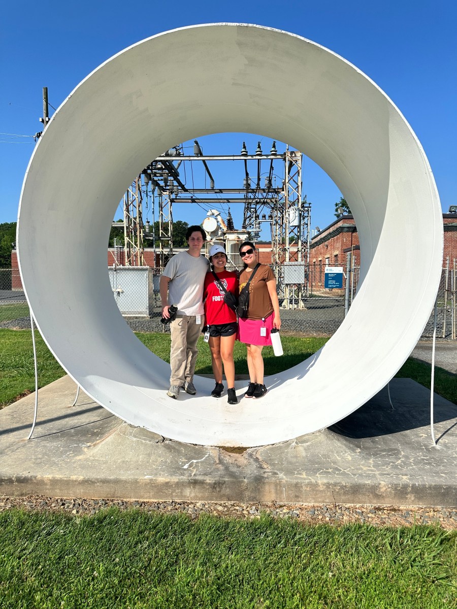 2 women and 1 man stand inside a very large section of white pipe with a grey metal utility power station hub in the background