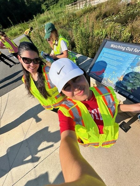 Selfie-photo of 2 student interns in yellow safety vests as they stand outdoors next to a tour sign