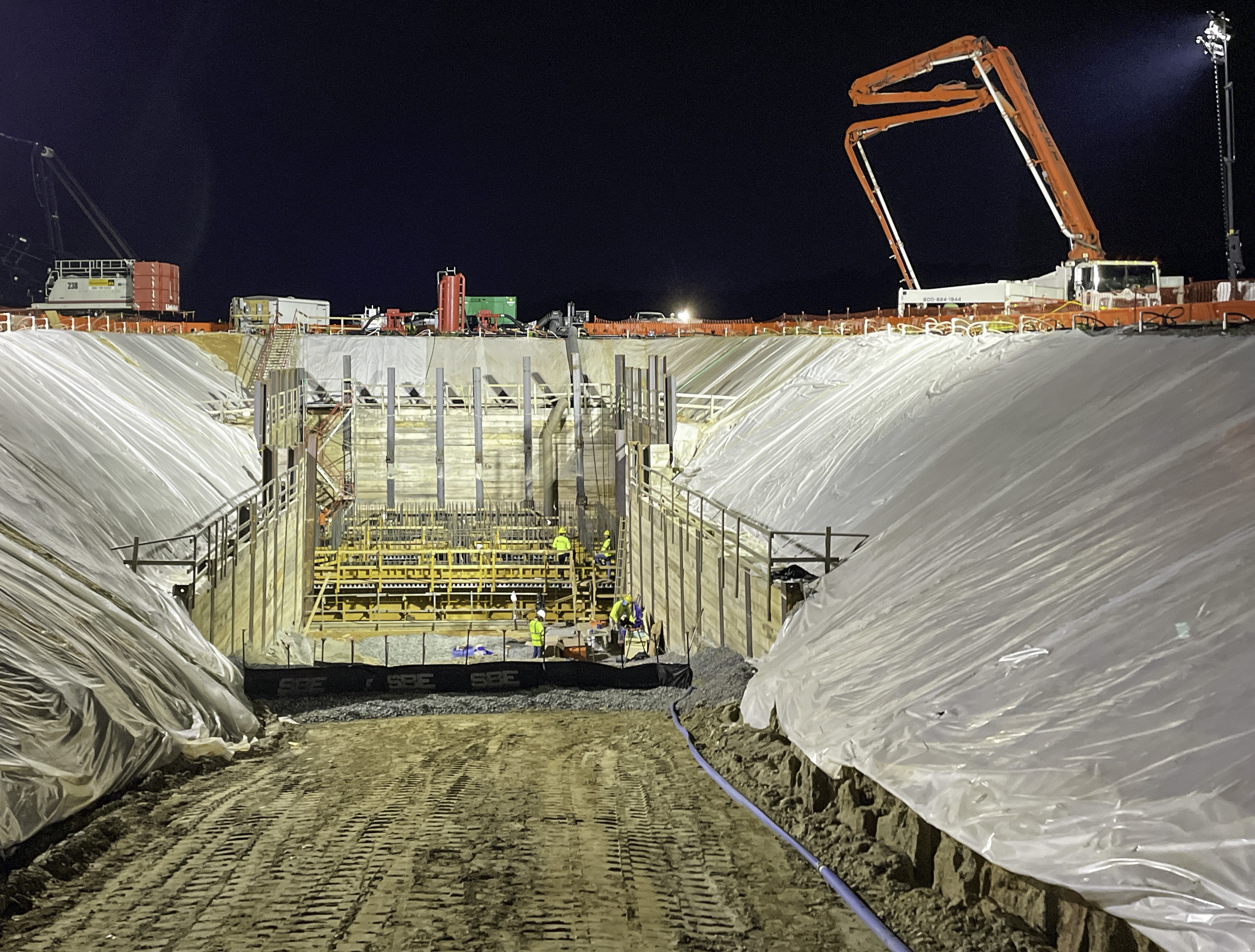 Utility crewmen in yellow safety gear build a pumping station at night at the base of a large wood-lined coffer dam; above and around them is earthen sloped ground shrouded with clear plastic sheeting, construction crew vehicles including a very large orange and white crane, and a giant light on a tower