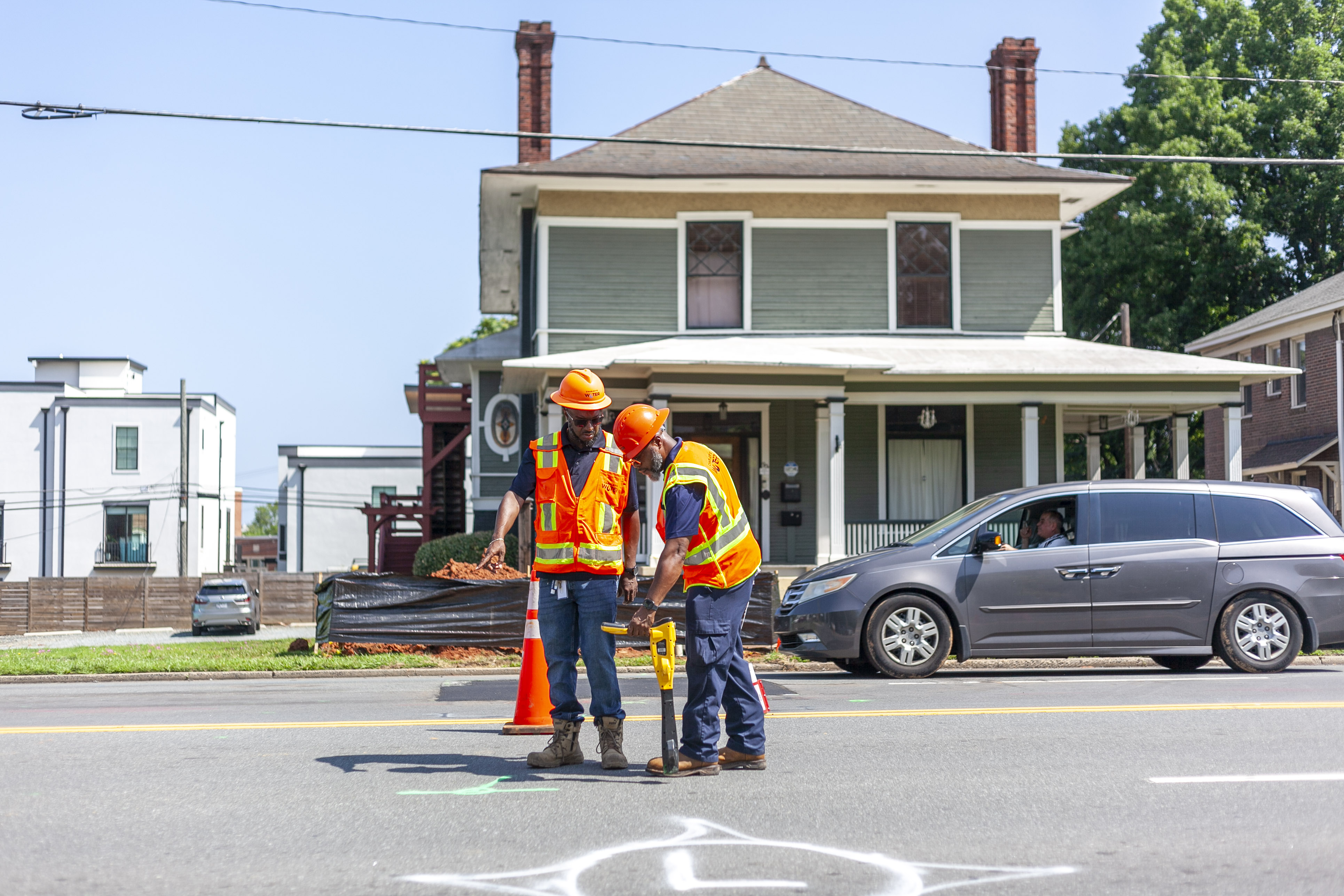2 utility crewmen wearing orange safety vests and orange hart hats use a yellow and black tool to mark a spot on the street in front of a sage green two-story house behind them