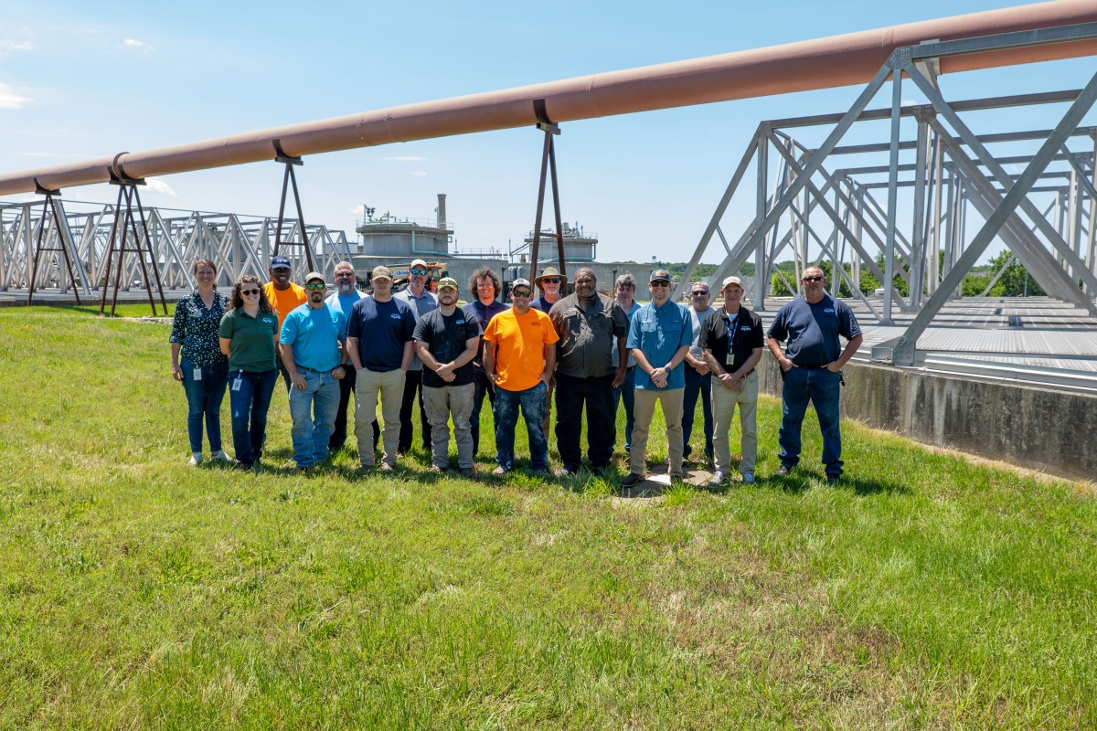 Over a dozen utility technicians pose on grass for a group shot in front of a water treatment plant; a large brown pipe runs behind and above them