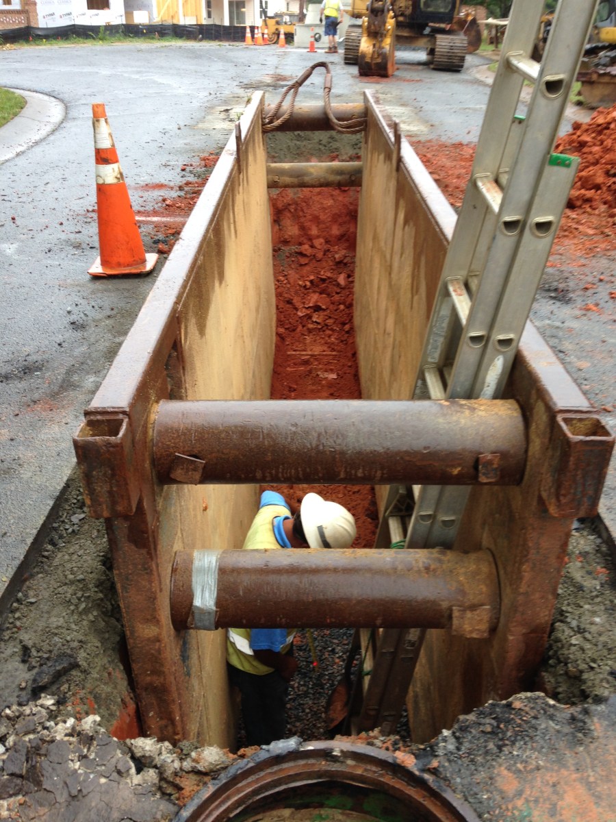 A deep trench cut into the terracotta red earth below a driveway is lined with a brown, metal septic chamber outfitted with re-enforcement rods; a utility crewman wearing yellow safety vest and white hard hat stands at the bottom with an aluminum grey ladder next to him