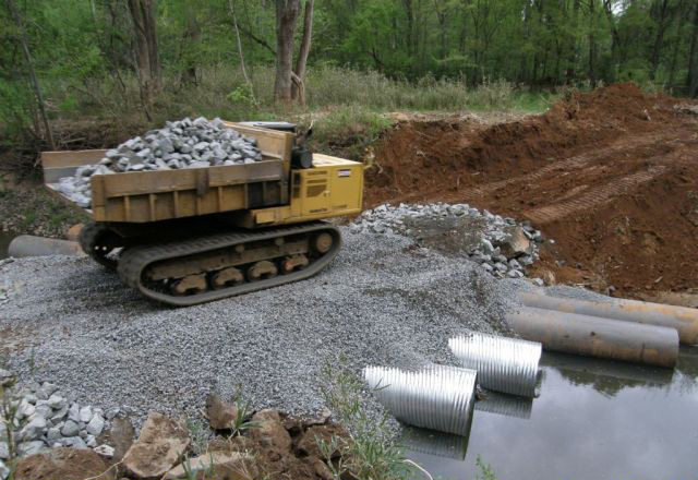 Large yellow, automated dump truck with caterpillar tracks filled with chunks of grey rock, moves from gravel path towards a dirt path overlooking 4 large metal run-off pipes below