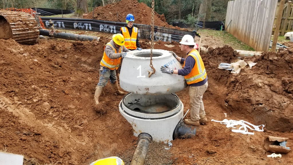 3 utility crewmen wearing orange safety vest and blue, yellow and white hard hats work with the help of a large chain to lower a large, grey concrete cap onto a large, grey concrete water junction chamber half-buried in the muddy ground