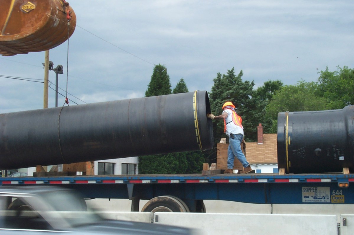 1 of 2 very large sections of black pipe is lifted off of a truck flatbed as crewman with orange safety vest and hard hat helps guide.