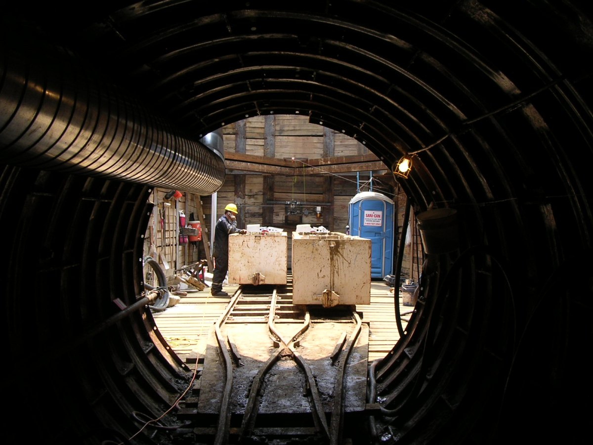 From the perspective of utility tunnel, view of a utility technician standing below ground, wearing a yellow hard hat reviewing plans via cellphone using a white metal cart as a table