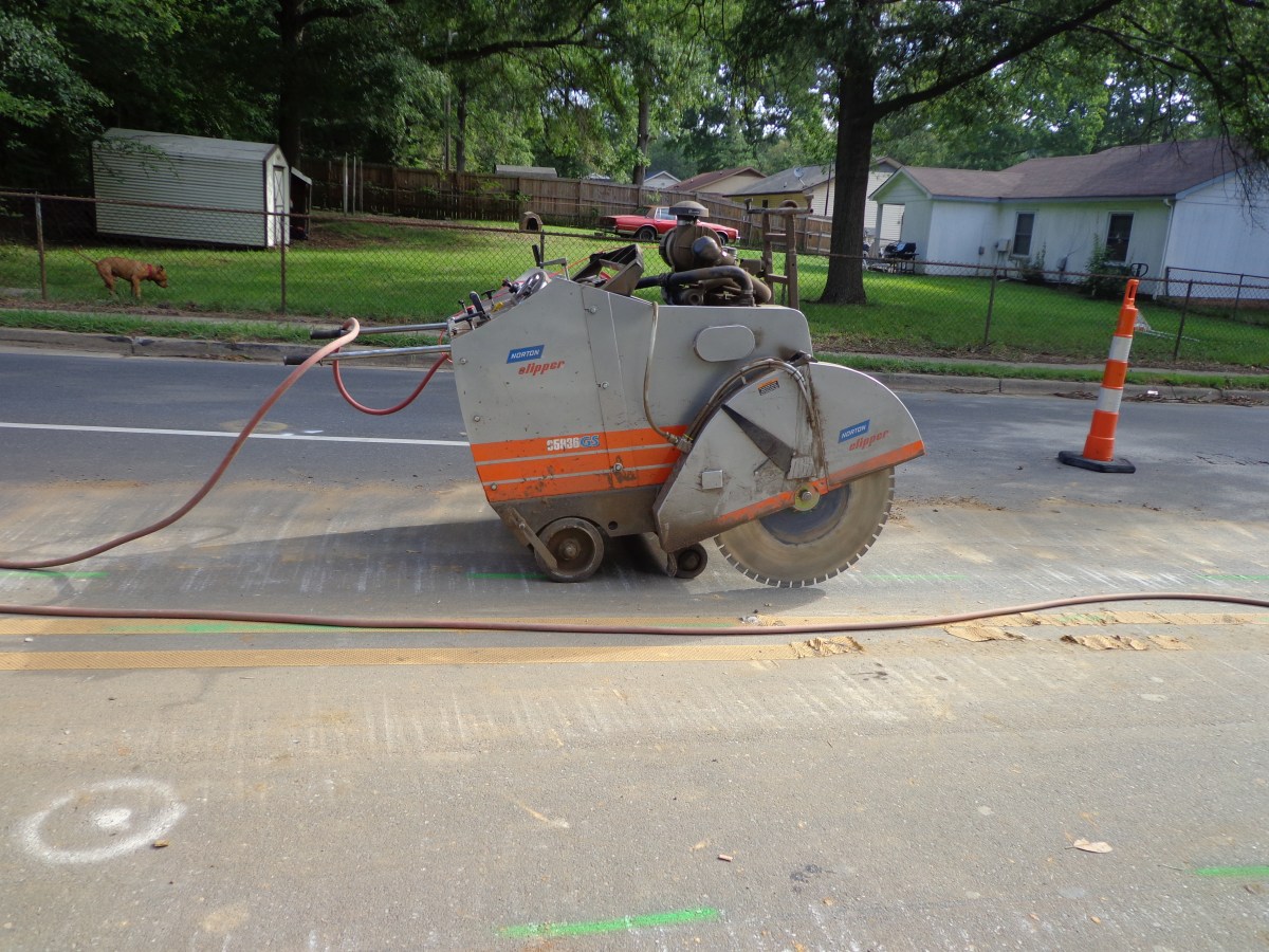 Grey and orange robotic macadam cutter with large circular blade sits on a residential street with an orange traffic cone