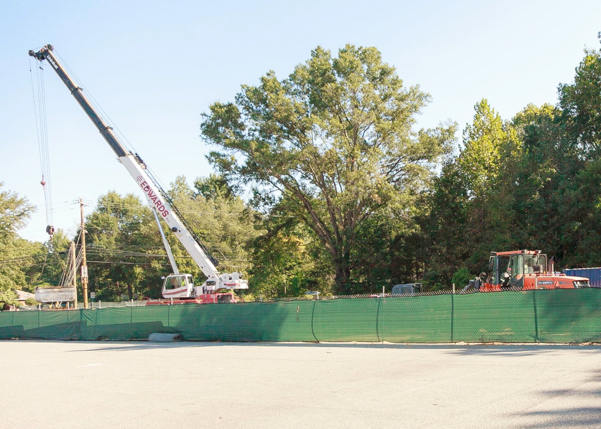 Large white crane with cable and tackle lowers a large block behind a green construction screen at a job site