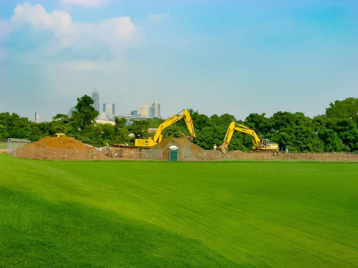 2 yellow excavators with their scoops move backfill dirt along a long pile of dirt next to a large expanse of bright green lawn; in the background is a cityscape against a blue sky and in the foreground, a dark green porta-potty.