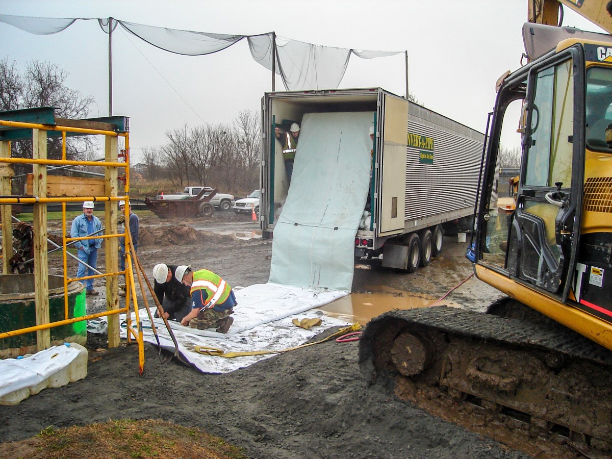 2 utility managers kneel on white material on muddy dirt by the back of a semi-truck and next to a yellow excavator's caterpillar tracks and yellow scaffolding