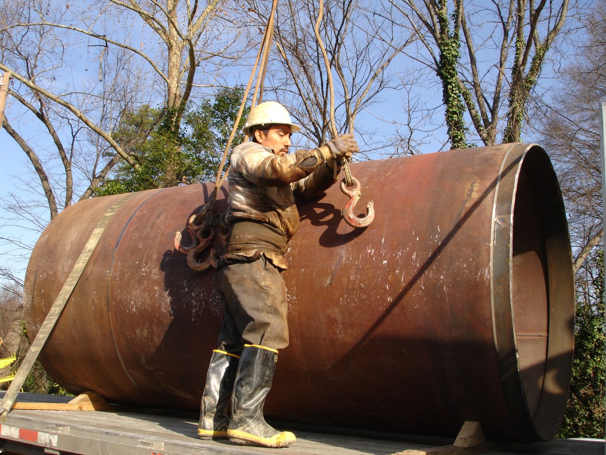 Technician in brown clothes and tan hard hat stands on a truck flat bed working to affix hooks onto very large section of brown, steel pipe