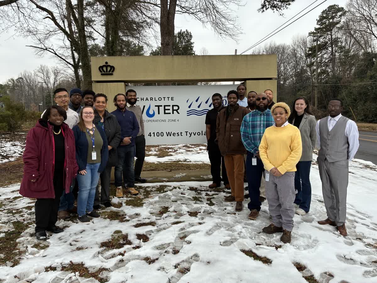 17 utility apprentices and staff pose outside in the snow in front of the Charlotte Water department's roadside sign