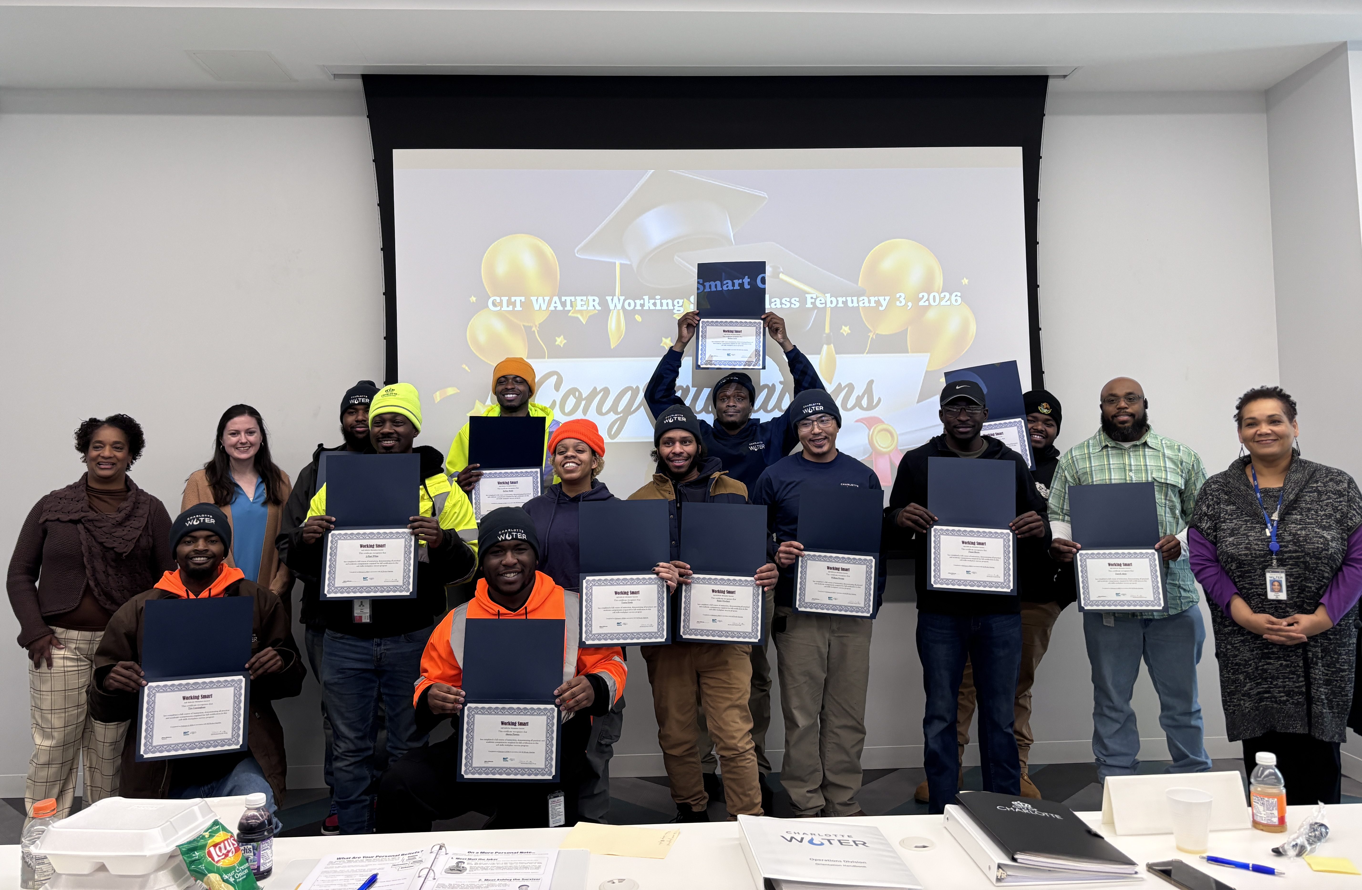 11 utility apprentices holding their certificates with 4 staff members line up for a group photo. Behind them there is a projector displaying "Congratulations"