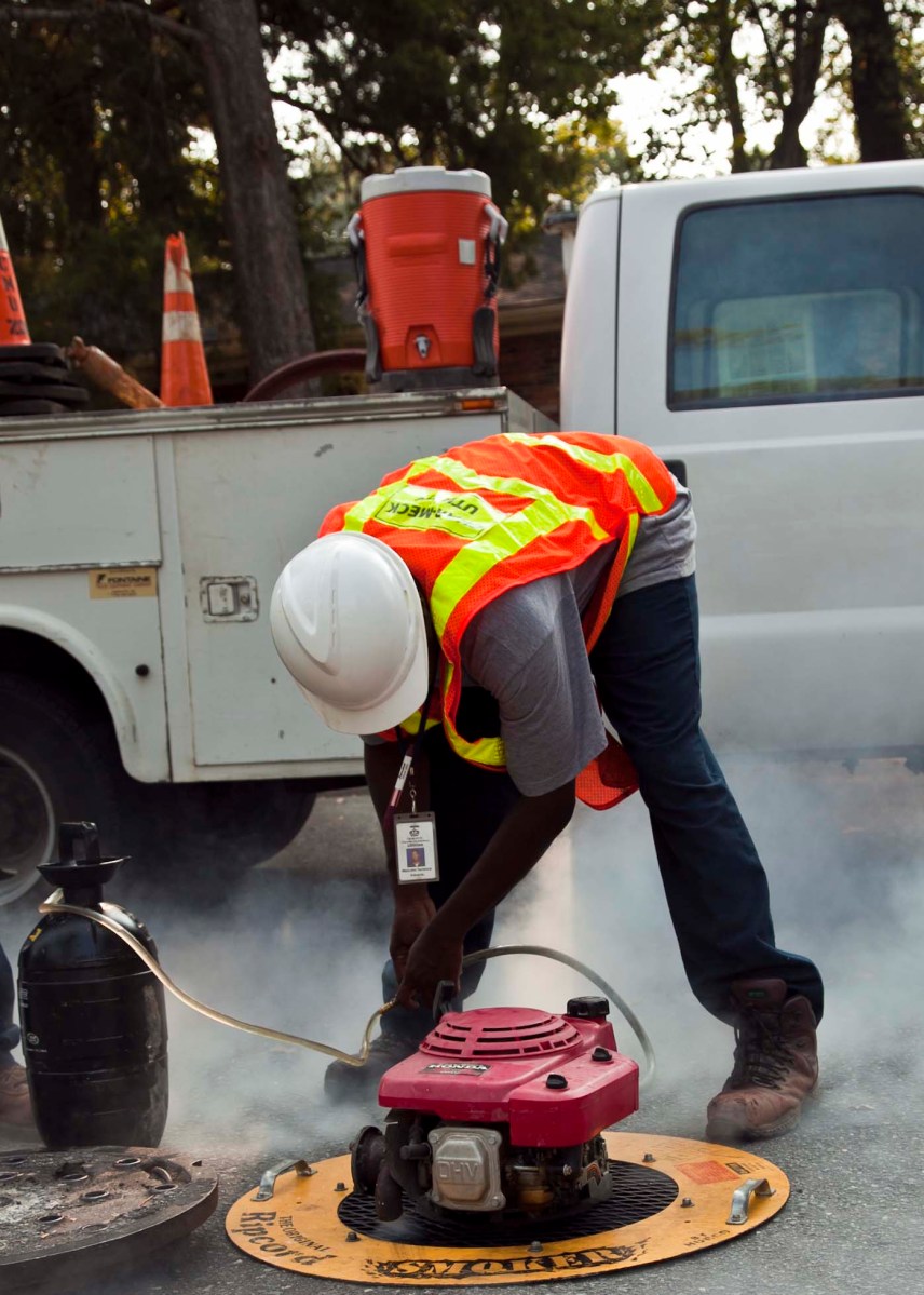 Utility worker wearing an orange safety vest and white hard hat bends down to examine a small, round red machine sitting on the payment