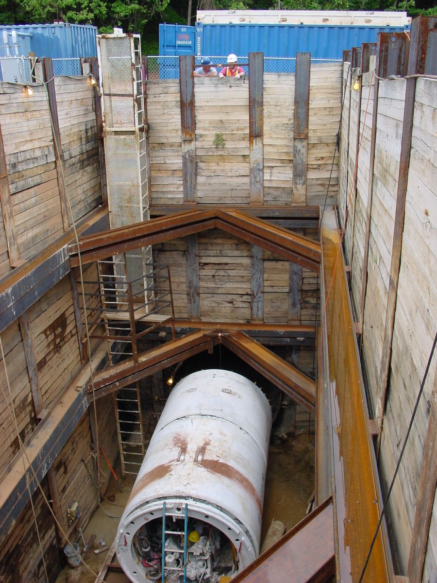 2 utility crewmembers with hard hats look down into a wood-lined, deep construction pit with a large white section of utility pipe at bottom