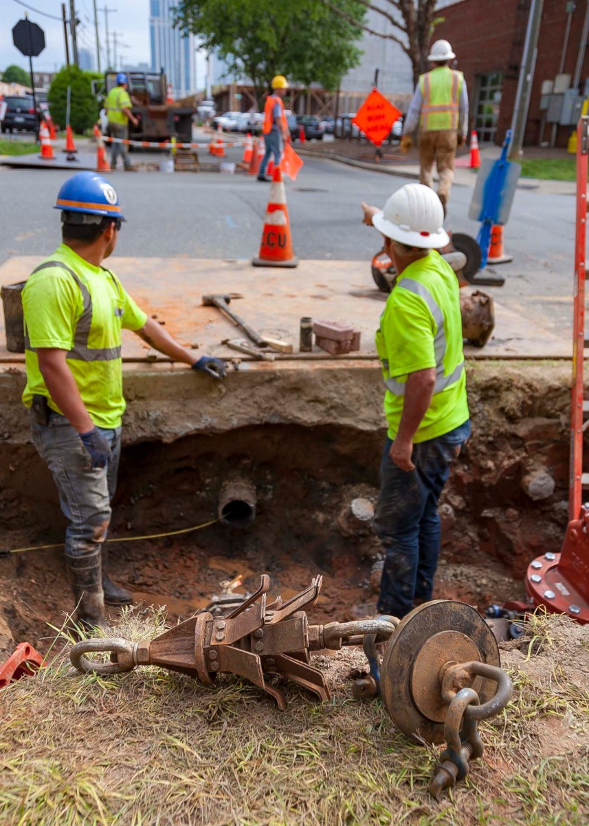 2 utility technicians with yellow safety vests and white and blue hard-hats stand in a street-side job site that is several feet below ground with a large piece of brown, metal piece of equipment next to them
