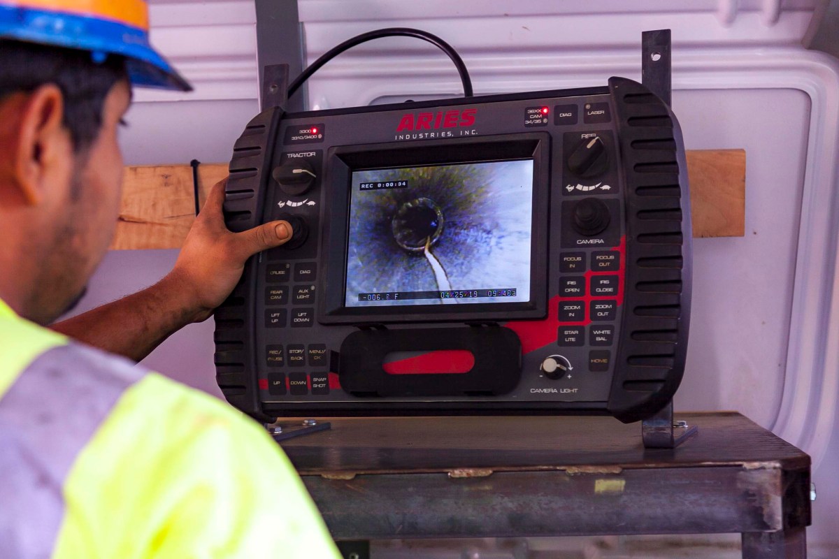 Utility technician operates a dark grey and red instrument panel with a video screen; he is controlling a camera that snakes its way through underground pipes