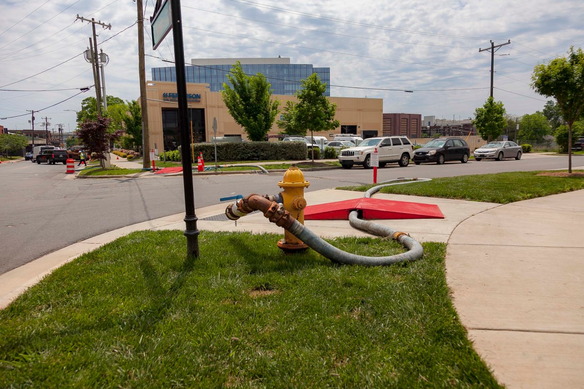 Yellow fire hydrant stands on a green, grassy, corner curb with grey hose hooked up to it; hose runs under red ramp to prevent pedestrian tripping as it snakes its way to the sewer drain streetside next to the curb