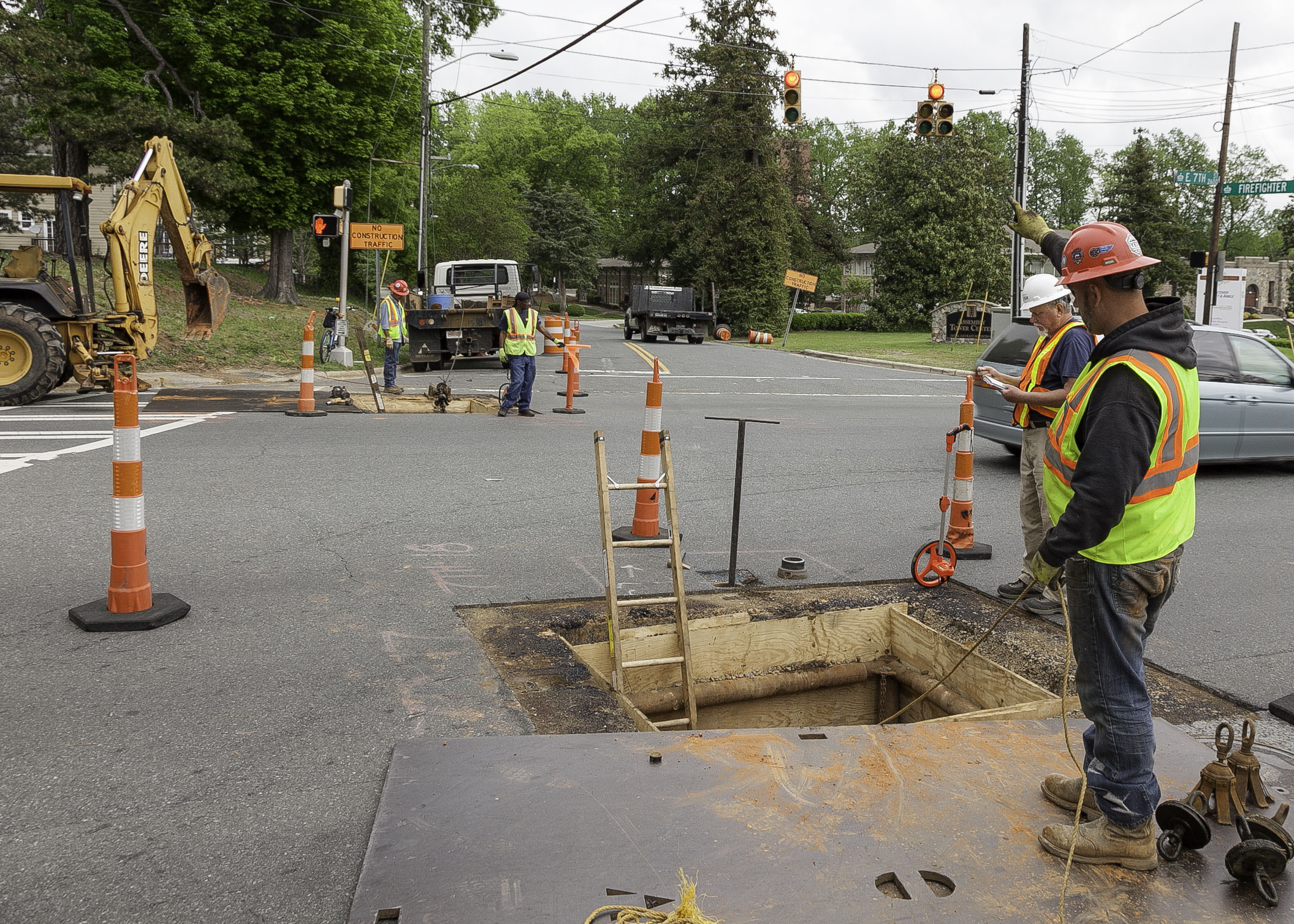 2 utility crewmen wearing yellow safety vest and hard hats examine a square hole and shaft that has been cut into a roadway, A ladder emerges from the shaft and is flagged with orange and white traffic cones