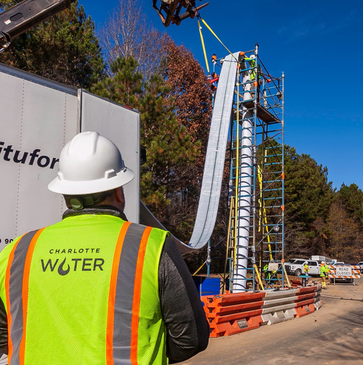 Utility worker wearing a yellow safety vest with the Charlotte Water logo watches an expandable fabric water pipe unrolled from the back of a white truck and lowered into the ground via tall scaffolding