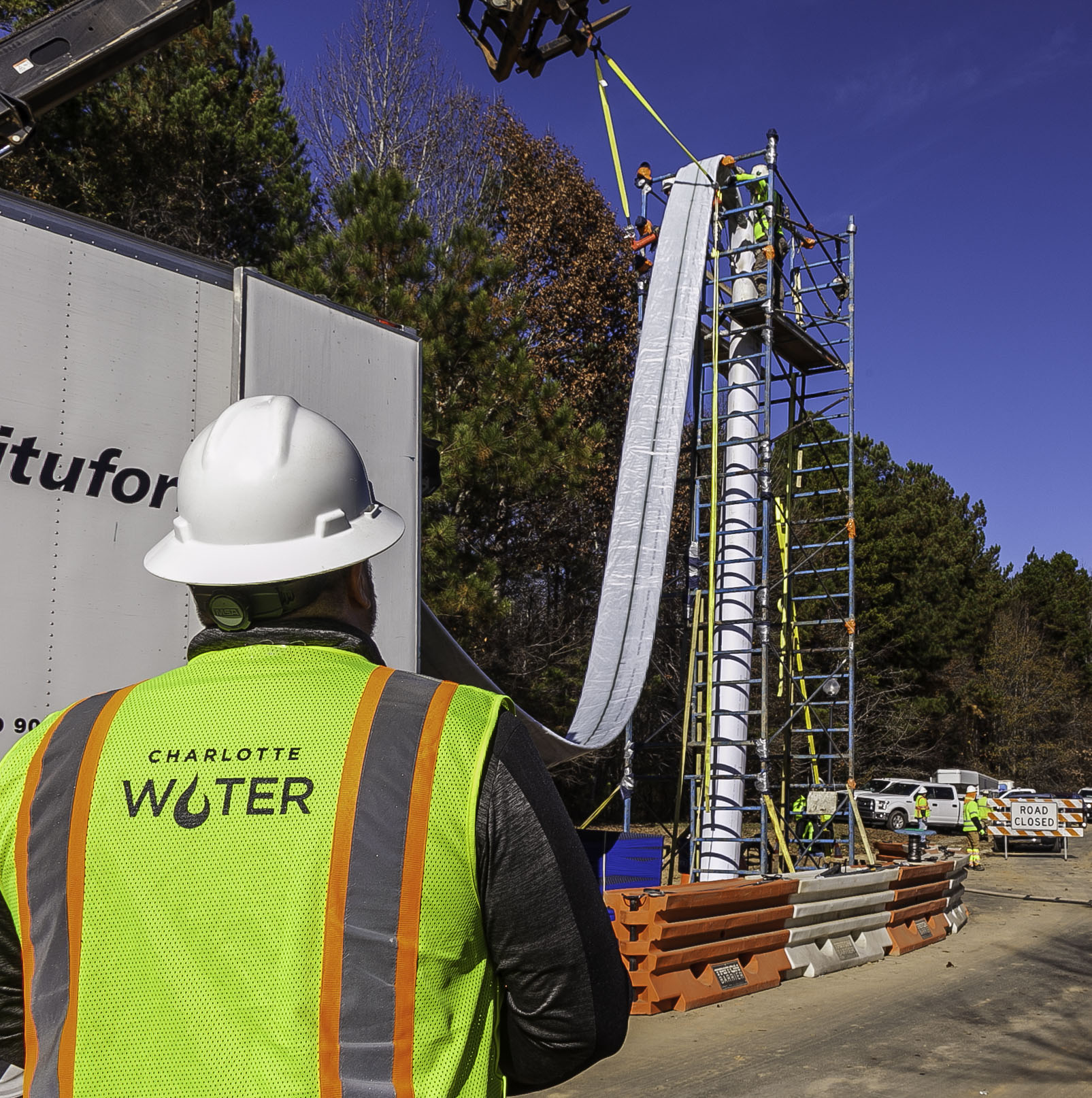 Utility worker wearing a yellow safety vest with the Charlotte Water logo watches an expandable fabric water pipe unrolled from the back of a white truck and lowered into the ground via tall scaffolding