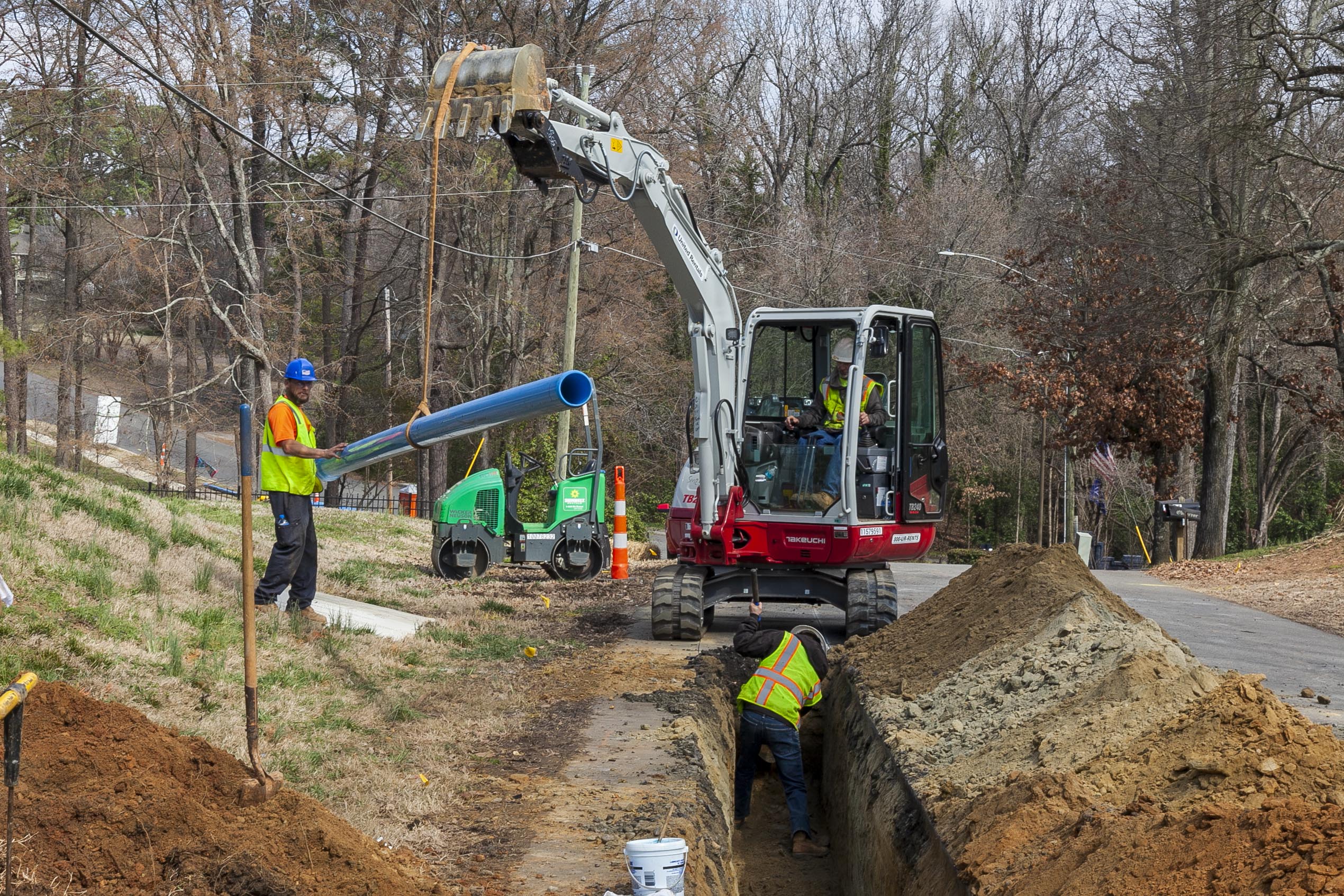 utility crewman wearing yellow safety vest and blue hard hat next to a grey backhoe helps lower a blue pvc pipe into a dirt trench