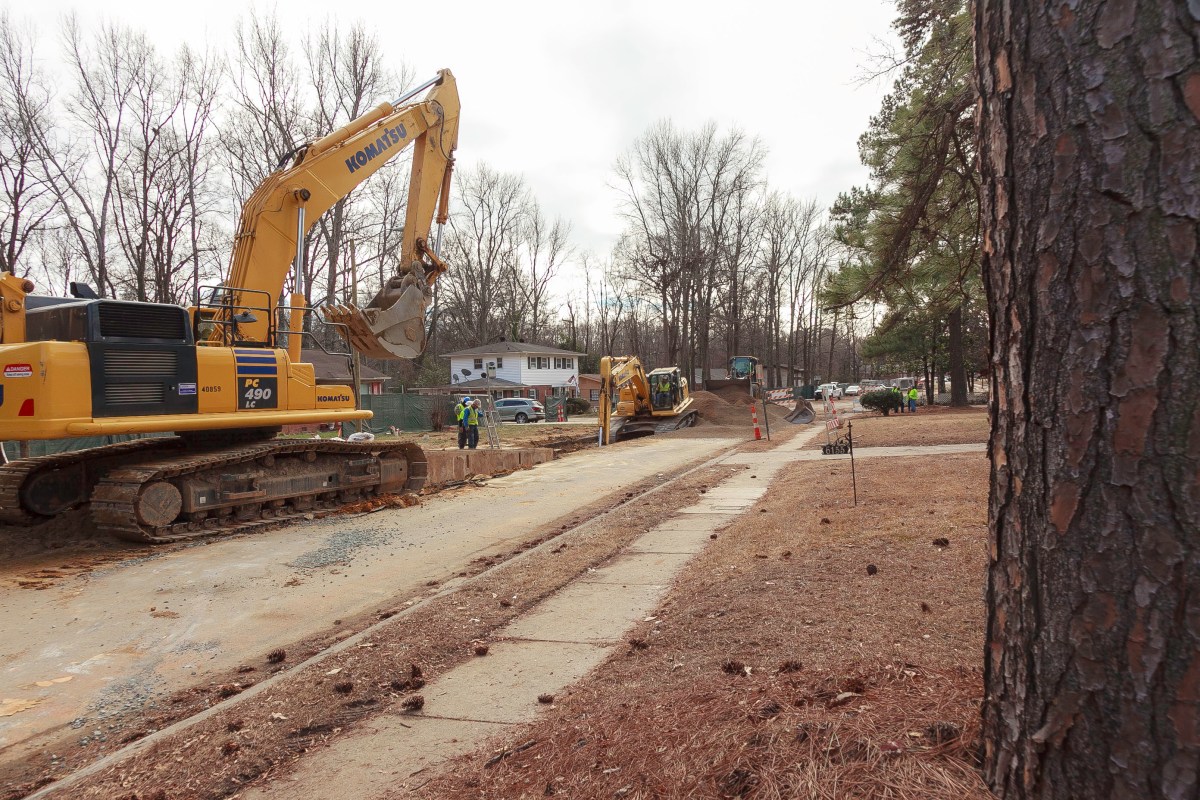 On a suburban street with leafless trees and several houses, 2 yellow, large excavators on caterpillar tracks guided by 3 technicians dig a long, deep trench of dirt