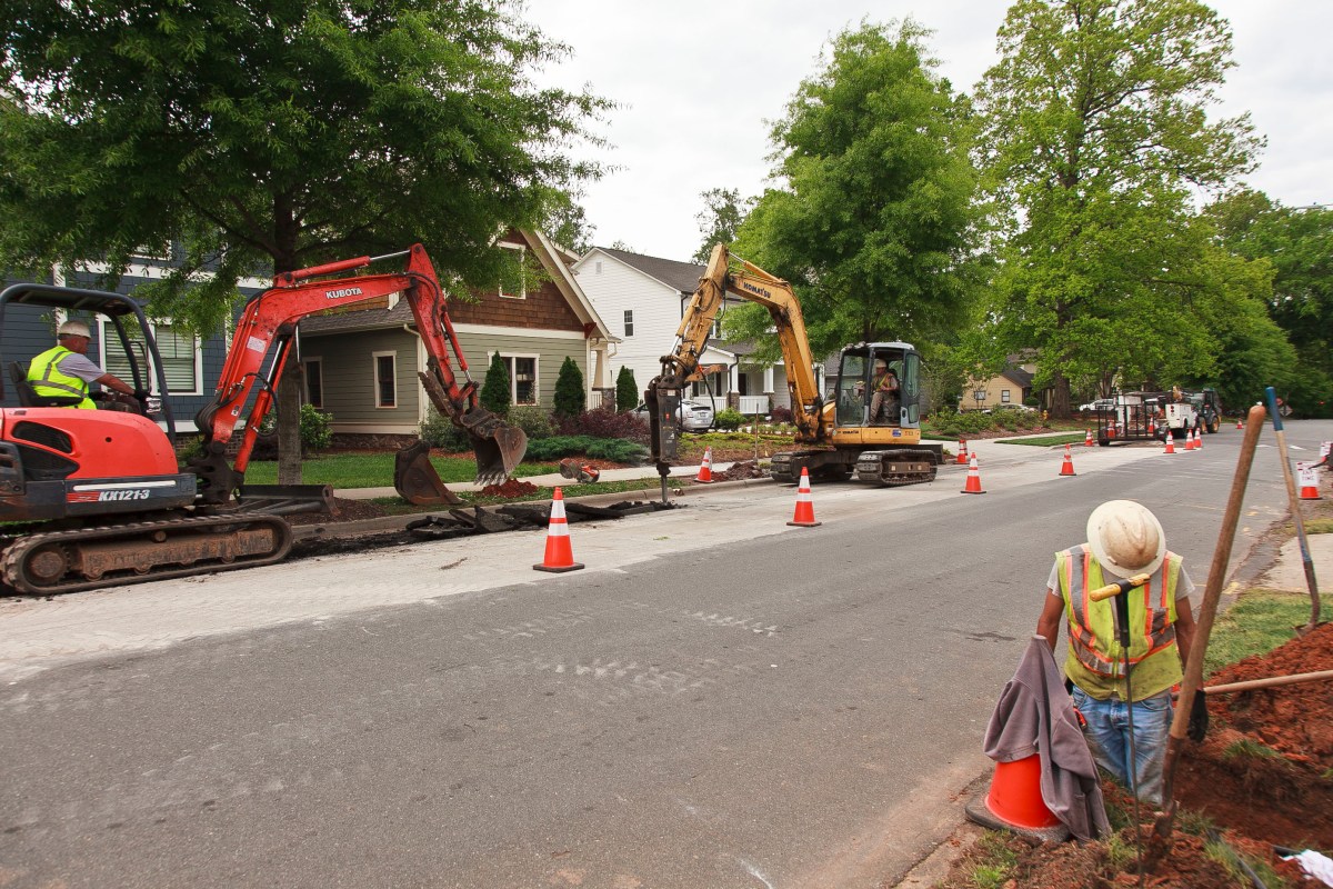 Utility technician with a hard hat examines a small trench curbside along a suburban street; behind him are a dozen orange traffic cones, a red excavator and a yellow backhoe with a pavement drill; both are driven with crew members with hard hats