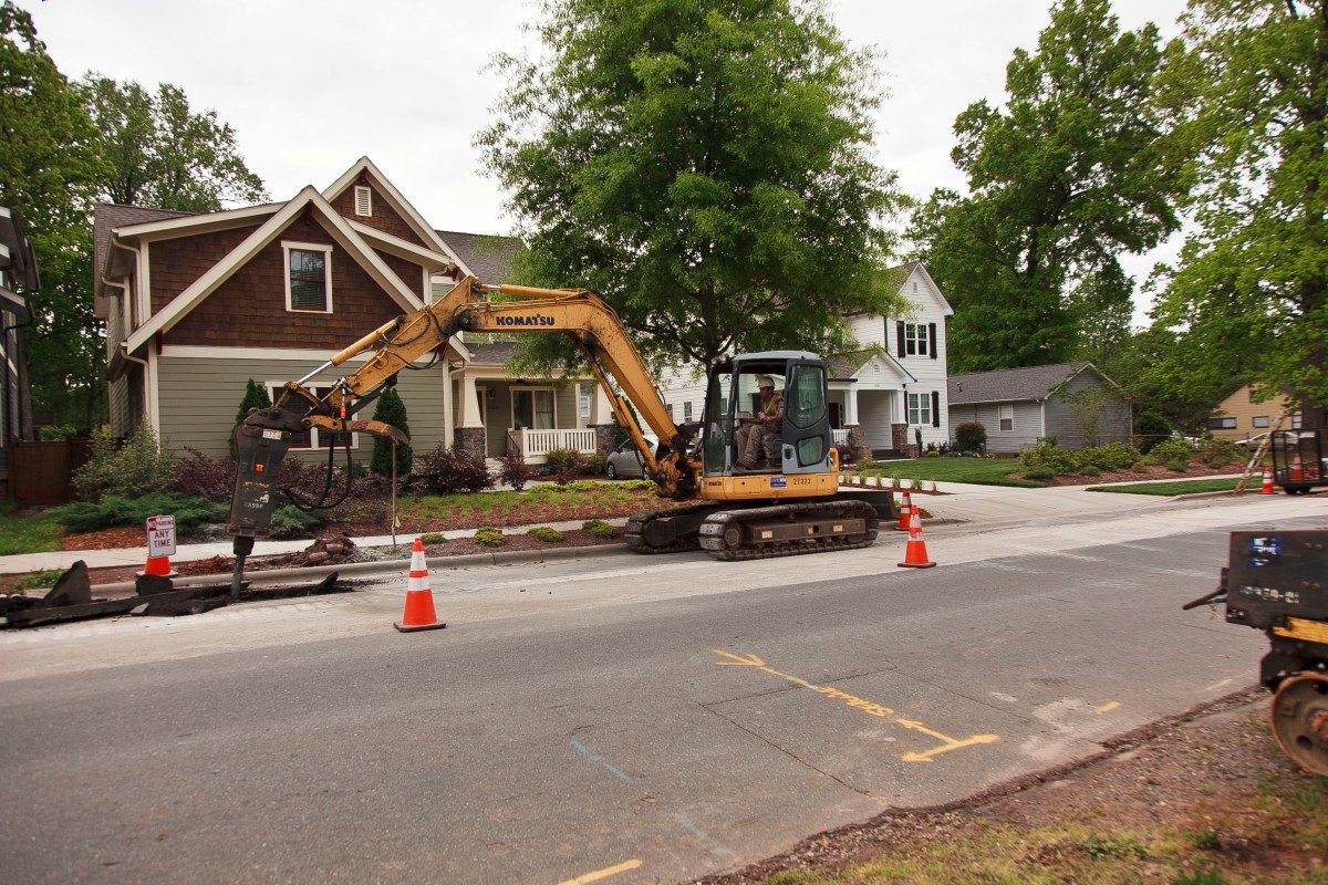 Yellow excavator with drill-head and caterpillar tracks cuts through the macadam on a suburban street blocked by orange traffic cones