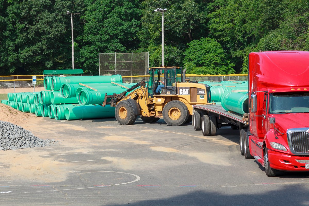 Dozens of sections of large light green pipes being stacked by a yellow CAT excavator next to a red flatbed truck