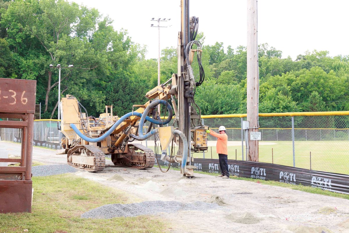 Yellow water drilling rig drills into dirt path next to a softball field