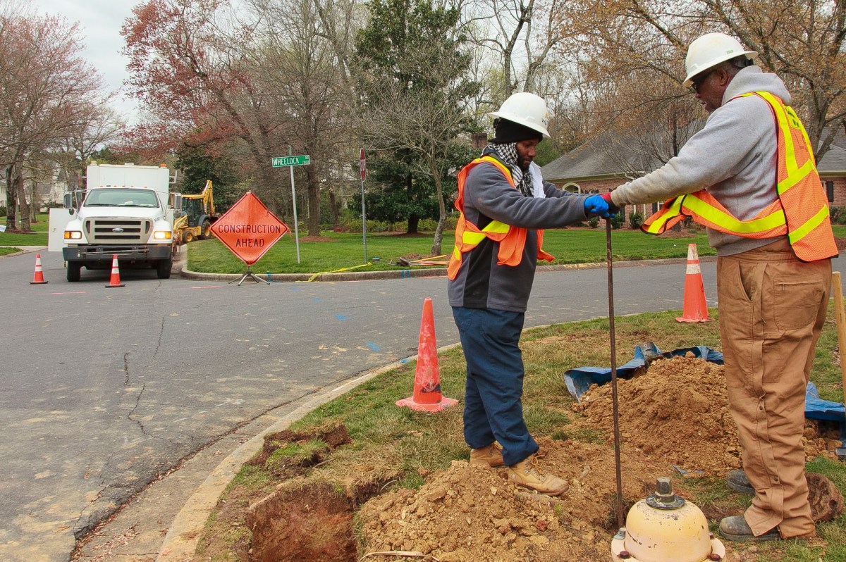2 utility technicians wearing orange safety vests and white hard hats stand near a yellow fire hydrant to turn a large metal pole called a key. They are standing on a grassy street corner in a suburban neighborhood with a white utility truck behind them