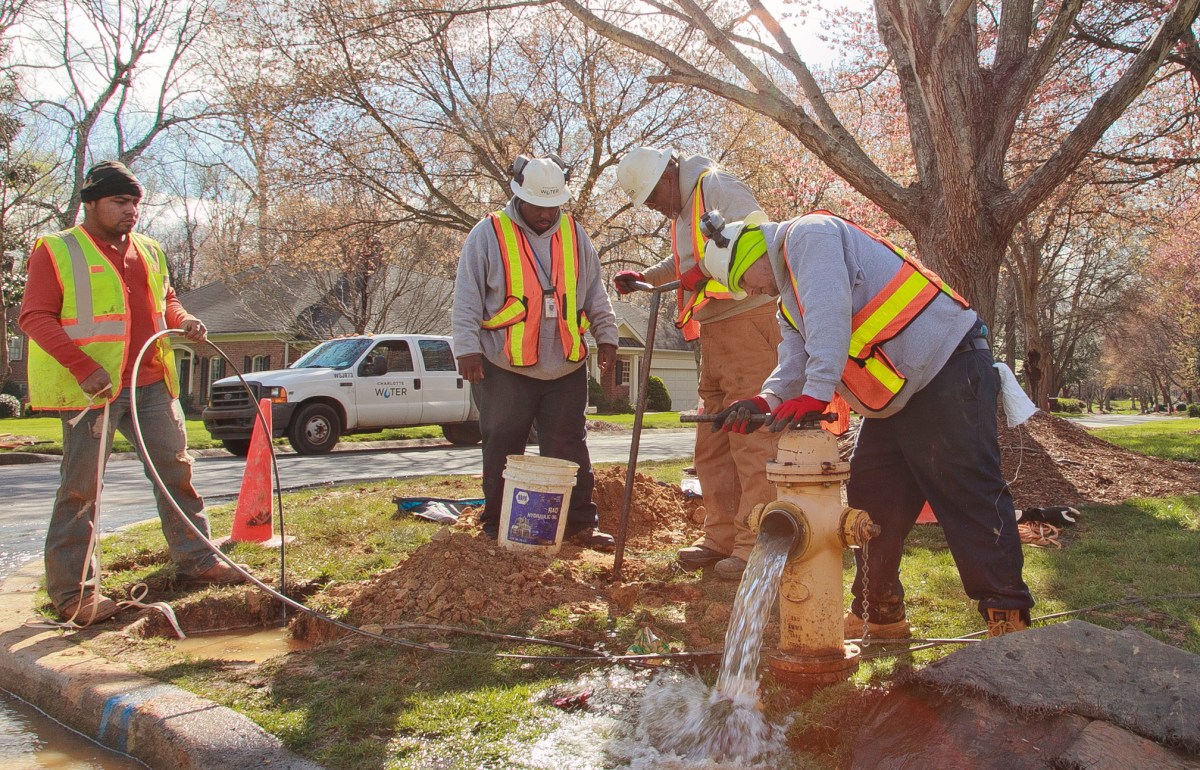 4 utility crewmen wearing yellow and orange safety vests and white hard hats test a yellow fire hydrant using a large wrench causing water to pour out of the hydrant and turning a mechanism in the ground with a large steel tool called a key