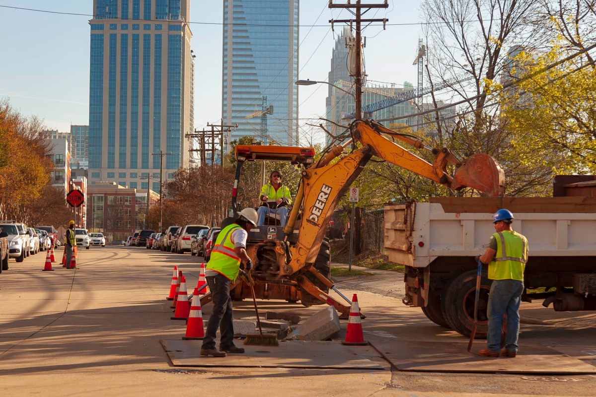 Utility technician in a yellow safety jacket drives a large, yellow John Deere backhoe excavating dirt from a trench on a street into the back of a white dumptruck; 2 additional utility technicians wearing yellow safety vests and white and blue hardhats stand nearby with push-broom, shovel and 5 orange traffic cones