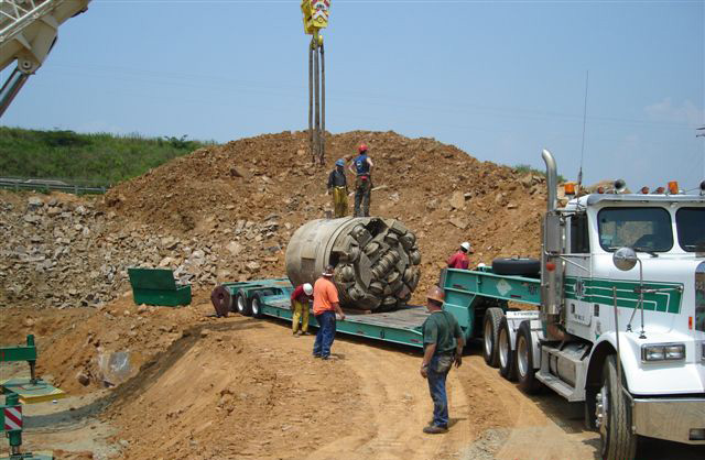 Section of brown, cylinder underground tunnel digger on a truck flatbed with 2 crewmen standing on top 4 others standing nearby; a large pile of excavated earth is behind them