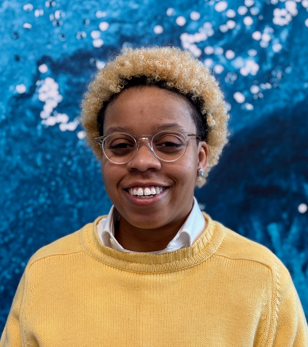 Profile photo of utility apprentice with curly blonde tipped dark hair, she is wearing glasses, earrings, a white collared shirt under a yellow sweater
