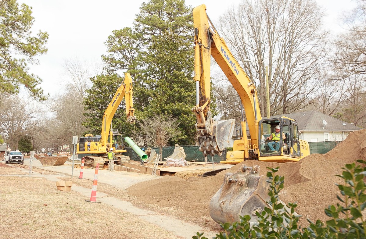Streetside in a suburban neighborhood, a large yellow backhoe with a crewman wearing yellow safety vest and white hard hat looks at pile of brown backfill dirt; nearby is a large yellow crane lowering a green section of pipe into a trench