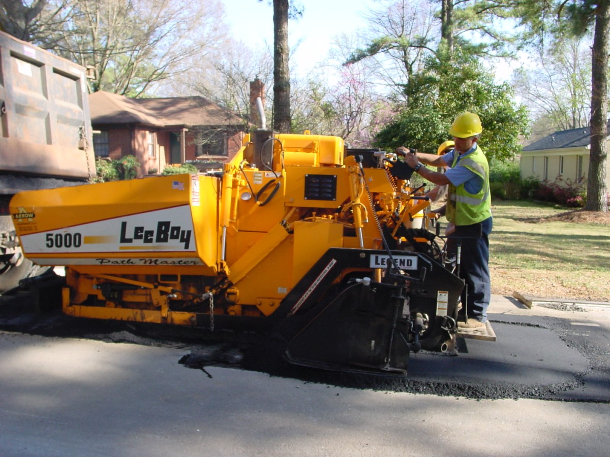 2 men wearing yellow yellow safety vests and hardhats ride on the back of a large, yellow LeeBoy Path Master paving machine watching as black macadam poured onto the roadbed is paved flat