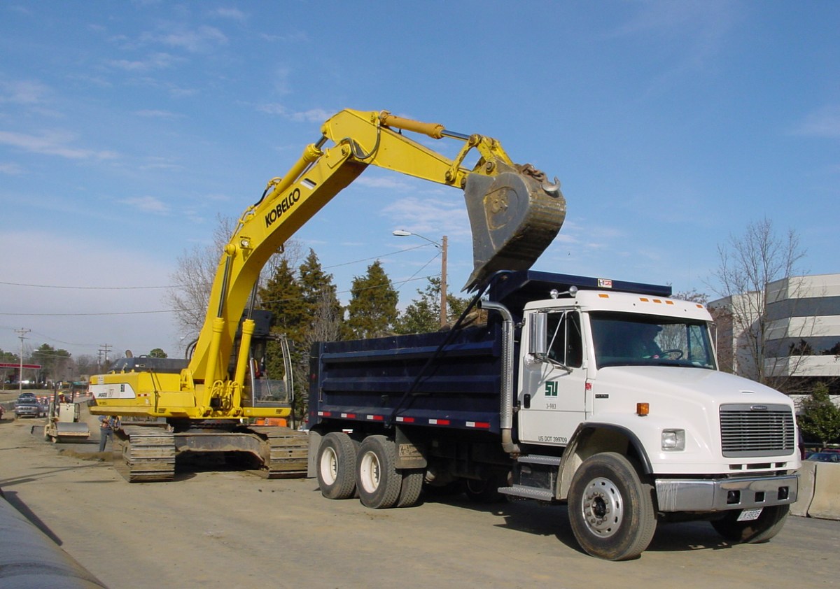 White and blue dump truck waits for the yellow backhoe behind it to finish filling it up. Behind both is a construction site in the roadway