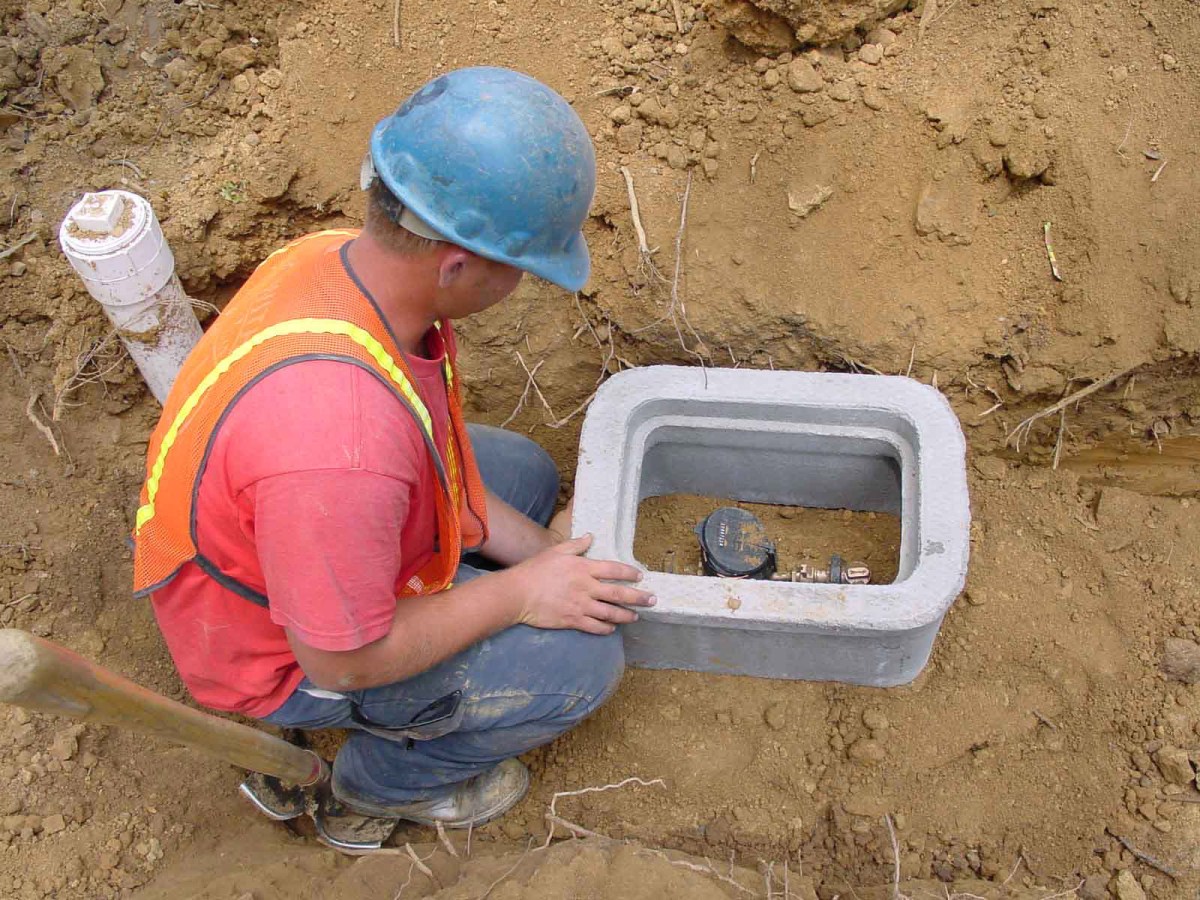 Utility technician with red shirt, orange safety vest and blue hard hat looks down at concrete meter box in the dirt with a black meter device installed inside