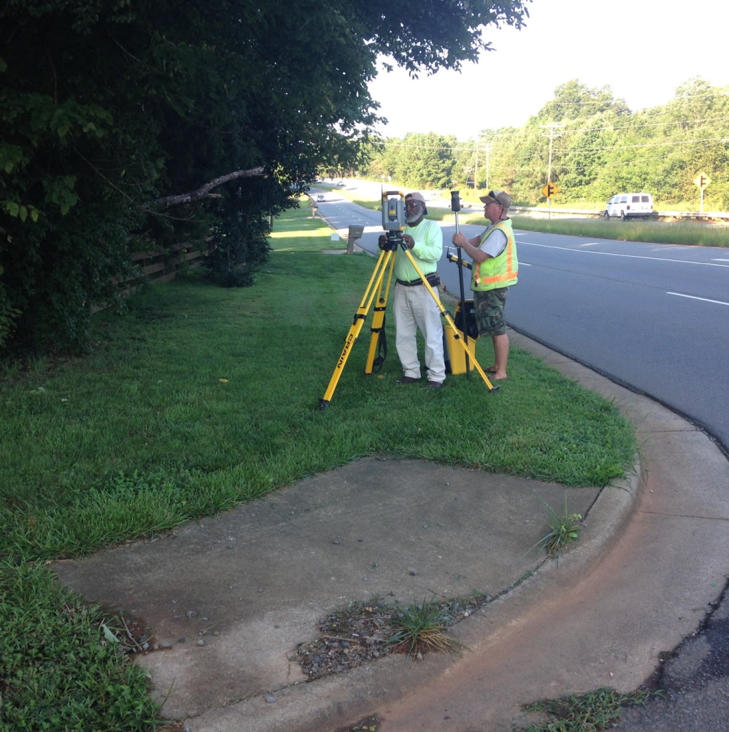 2 surveyors wearing yellow safety vests stand next to a yellow survey tripod and a gps indicator on a pole