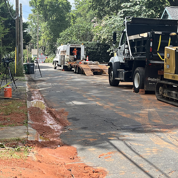 2 trucks parked along the side of a residential tree lined street; across the road is dirt and mud from a leak