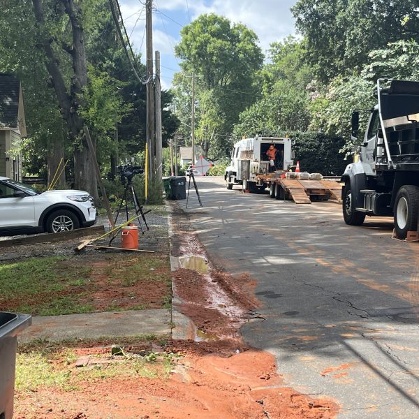 2 trucks parked along the side of a residential tree lined street; across the road is dirt and mud from a leak