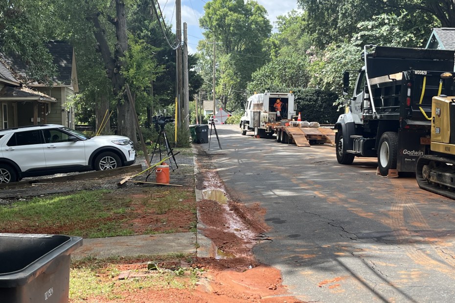 2 trucks parked along the side of a residential tree lined street; across the road is dirt and mud from a leak